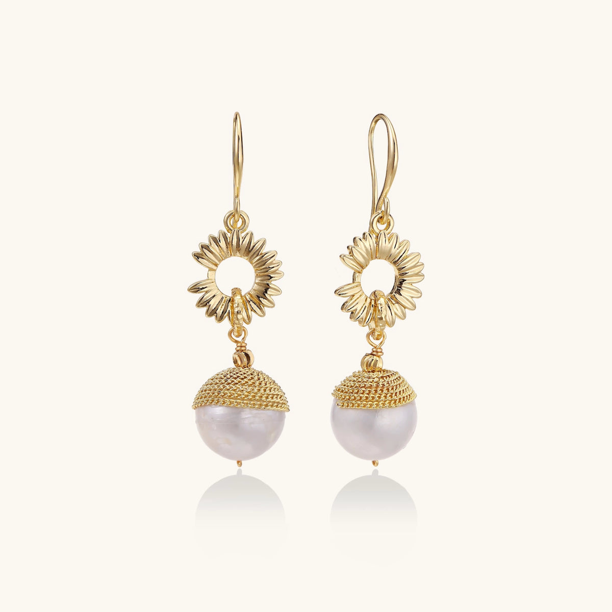Gold sunburst charm earrings with baroque pearl drops on French hooks against cream background