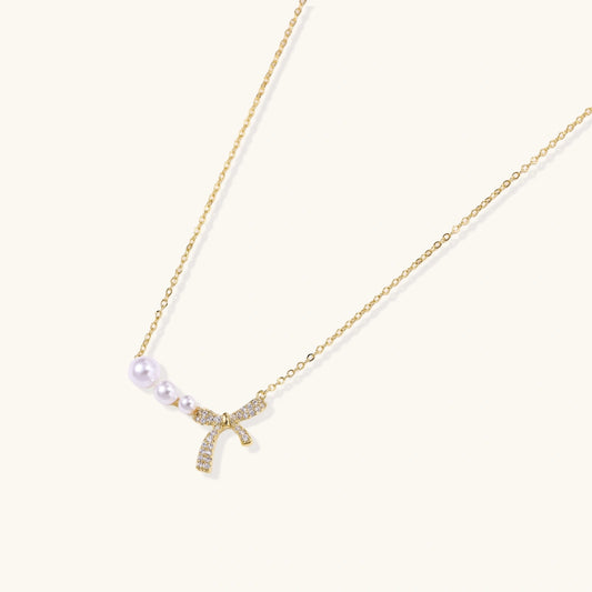 Delicate gold necklace with three white pearls and crystal-embellished bow pendant on cream background